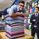 A-wide-humorous-scene-in-a-retail-store.-An-employee-with-a-sly-smile-is-seen-stacking-a-teetering-pile-of-neatly-folded-shirts-on-the-very-edge-of-